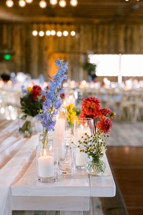 Blue delphinium, coral dahlia, yellow freeisa, feverfew chamomile bud vase and pillar candle centerpiece Wild Flower Bud Vase Centerpiece, Wildflower Bud Vase Centerpieces, Wildflower Candle Centerpiece, Floating Candles With Bud Vases, Delphinium Centerpiece Wedding, Bud Vase And Floating Candle Centerpiece, Pillar Candle And Bud Vase Centerpiece, Dahlia Bud Vase Centerpiece, Wildflower And Candle Centerpieces