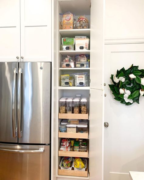 This vibrant kitchen features a large stainless steel refrigerator and bright white cabinets adorned with a leafy wreath. A narrow pantry is built into the wall and displays rows of clear bins with neat labels. Turn a small space in your kitchen into a pantry with stackable bins and canisters, and use labels to keep everything tidy. Small Pantry For Small Kitchen, Pantry In The Wall, Narrow Larder Cupboard, Narrow Space Next To Refrigerator, Fridge With Pantry Cabinet, How To Add Pantry To Small Kitchen, Pantry In Open Concept Kitchen, Small Pantry With Fridge Design Ideas, Narrow Pantry Next To Refrigerator