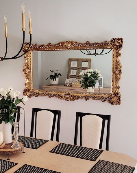 Incorporating gold ornate mirrors in your home decor in a refreshed way. This one is uses in the dining room with light wood table and black chairs. A ceramic vase with white roses makes for a simple timeless centerpiece. Ornate Gold Mirror Living Room, Gold Mirror Above Couch, Mirror Above Dining Table, Gold Mirror Living Room, Mirror Over Couch, Mirror Above Couch, Ornate Gold Mirror, Dining Area Ideas, Gold Ornate Mirror