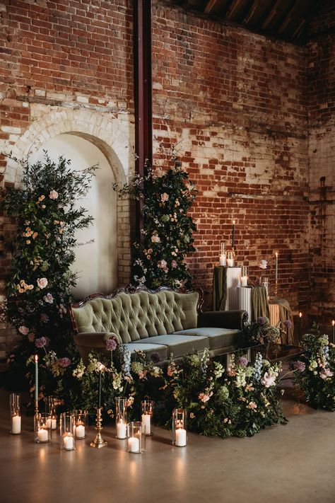 This shows a barn wedding lounge set up. The focus is on a green velvet three seater louis-style sofa. The sofa is surrounded by large flower arrangements and differently sized hurricane lanterns with pillar candles. Next to the sofa are also three ribbed round plinths, two white one wooden. On the plinths are flowers, lanterns and candles. Wedding Portrait Lounge, Portrait Lounge Wedding, Photo Lounge Wedding, Wedding Lounge Seating Indoor, Wedding Photo Area, Lounge Area Wedding, Photo Lounge, Wedding Lounge Seating, Wedding Lounge Furniture