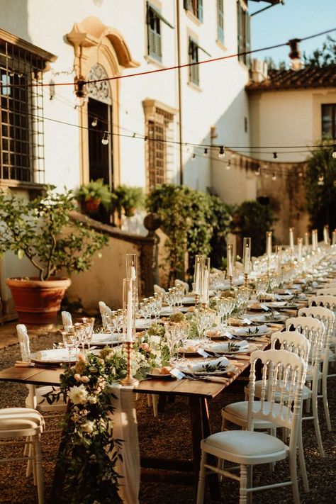 Wedding reception with a beautiful family style long wooden table, white romantic chairs, table runner made of eucalyptus, tea roses and white garden flowers, white taper candles in golden candelholders and vintage table settings under the string lights at a beautiful Italian garden of the 17th century Tuscan villa. White Chairs Wedding, Vintage Table Settings, White Garden Flowers, Wedding Flower Table Decorations, White Taper Candles, Family Style Weddings, White Flowers Garden, Family Style Table, Long Table Wedding