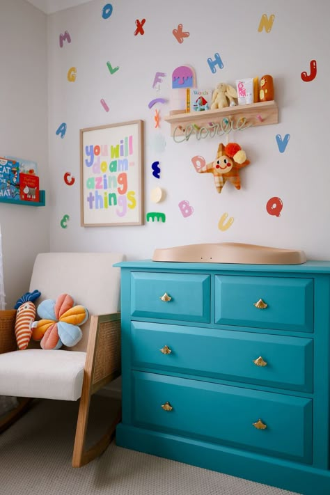 Teal blue dresser with changing tabel on top, white modern arm chair in corner, colorful letters stuck in non pattern on wall