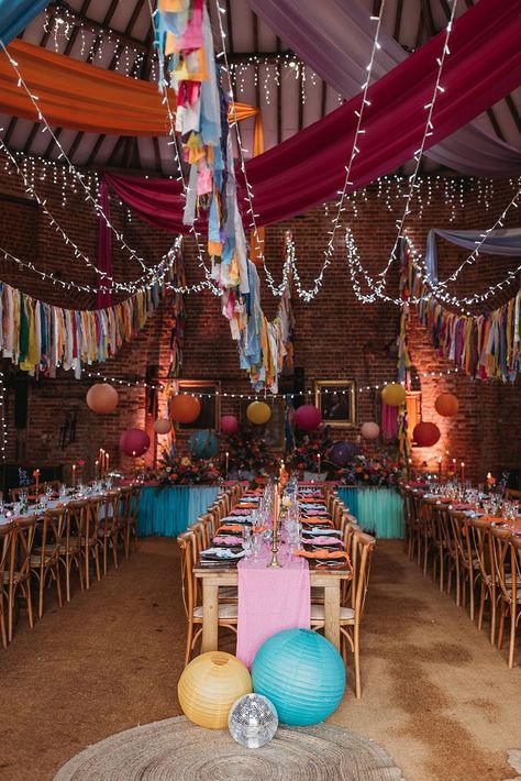 This shows a colourful wedding set up with three rectangular rows of dining tables. The focus is on the middle row of wooden tables. On the table is a pink table runner across the middle of the table. There are also black plates paired with many different coloured napkins. On the table are glassware and glass vases with flowers in them. There are also brass candlesticks with colourful candles.Above the rows of tables are colourful bunting. At the base of the table are round paper lanterns. Paper Lanterns Wedding, Wedding Setup, Wedding Dining, Dinner Setting, Vibrant Wedding, Asheville Wedding, Picnic Wedding, Wedding Venue Decorations, Wedding Set Up