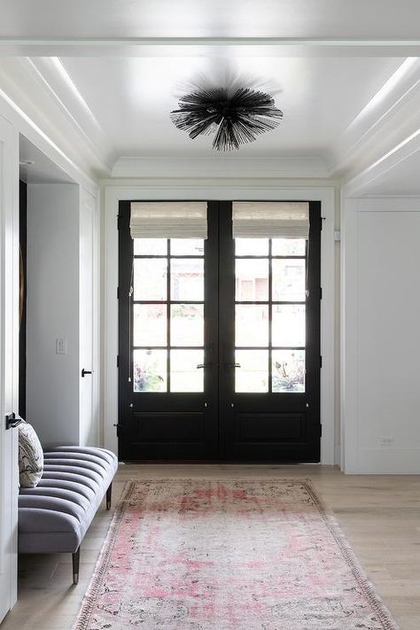 A red and gray vintage rug sits beneath a black sea urchin flush mount fitted to a tray ceiling in front of black double front doors boasting glass panels and white roman shades. Black Sea Urchin, Cottage Foyer, White Roman Shades, Door Shades, Rustic Wood Floors, Kate Marker Interiors, Double Glass Doors, Black Front Doors, Double Front Doors