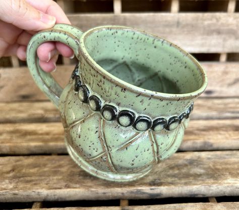 This 18 oz hand thrown and altered mug was handmade on my pottery wheel in Hubert, NC. After I threw the mug shape on my wheel with speckled brown stoneware clay I let it dry for a day and then altered the shape of the belly by pressing lines in it from the outside and pushing out on the inside. I attached a necklace of clay balls  with dark clay around it the neck of the mug. It has a strong handle that comfortably fits 3 fingers. The glaze is a nice sage green. It has a very sturdy base and is perfect for warming hands around its round belly!  Dishwasher, oven and microwave safe.  4 1/4 inches wide excluding the handle, and 3 3/4 inches tall. It holds 18 oz. of your favorite beverage. Carved Mug, Carved Pottery, Barbed Wire Art, Pottery Necklace, Mug Pottery, Pottery Glazes, Barbed Wire, Pottery Wheel, Stoneware Mugs