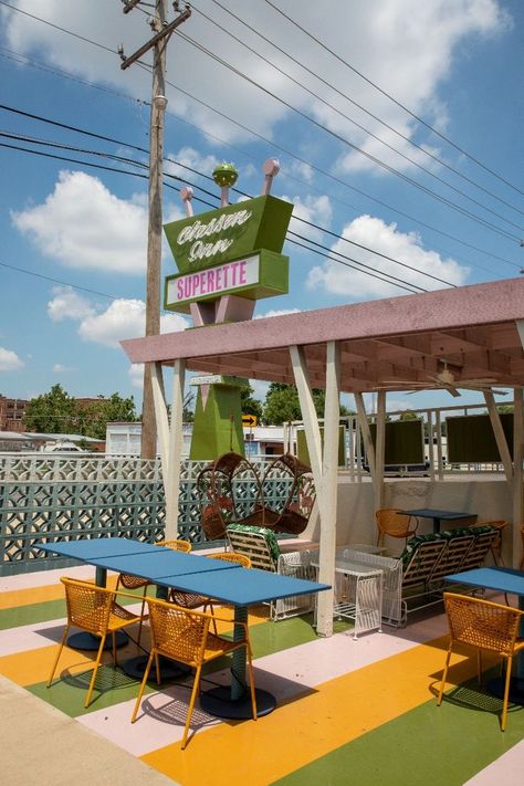 A five-photo carousel. First: a bed with a yellow spread and the wall behind it has a palm tree design with a pair of large, teal colored lips. Second: an orange, pink and green striped mural shaped like a rainbow in a guestroom. Third: the vibrant Suprette with many colors, a blue couch, pink chairs and small pieces of decor. Fourth: looking out the door from a guest room toward teal bikes under a staircase. Fifth: The patio with yellow chairs, blue tables and the hotel sign in the background. The Dive Motel, Mid Century Motel, 1960s Motel, Motel Design, Boutique Motel, Googie Design, Oasis Springs, Googie Architecture, Retro Motel