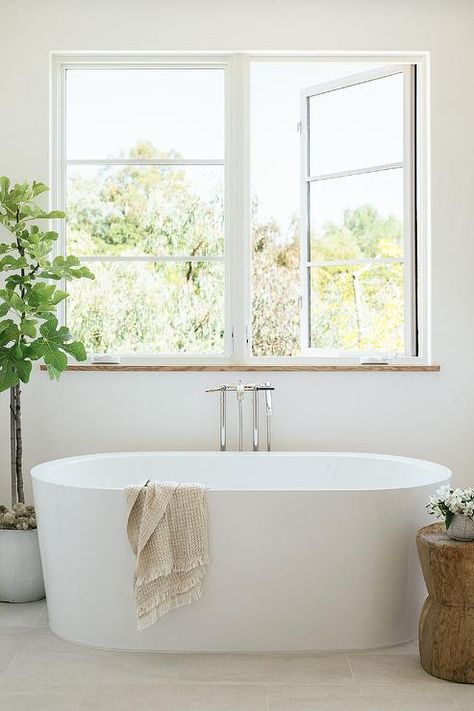 Bathroom features modern oval white bathtub with modern floor mount tub filler under windows, salvaged wood accent table and a potted faux tree. Bath Under Window, Concrete Bathtub, Contemporary Craftsman, Cottage Bathroom, Wood Accent Table, Transitional Bathroom, Bathroom Windows, Design Del Prodotto, Bathroom Renos