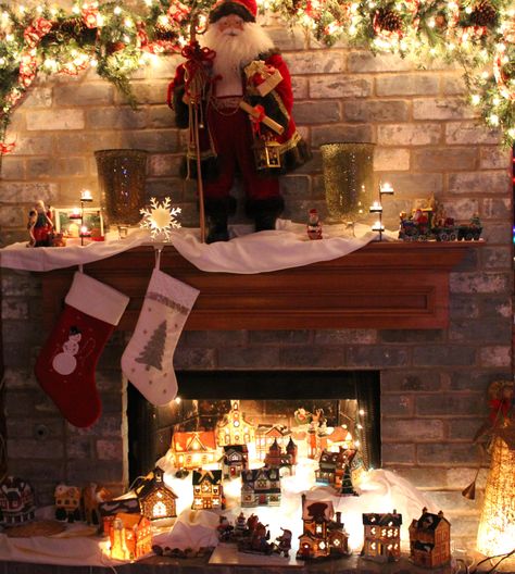 Our sister-in-law's mantel and village scene in their fireplace. She used Christmas lights under white satin along with some glass blocks to make a snow-side hill for the village. Fireplace Christmas Village, Christmas Village In Fireplace, Below Tv Christmas Village, Santa’s Village Decoration, Christmas Village Water, Christmas Village Sets, Lemax Christmas Village 2022, Village Ideas, Diy Christmas Village