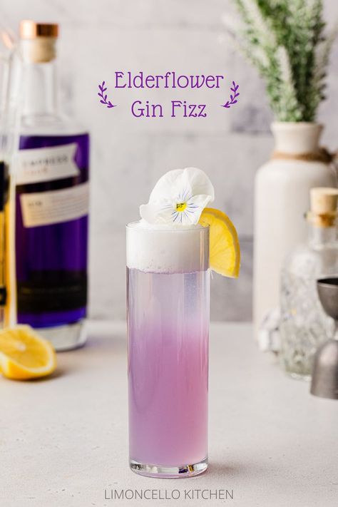 Side view of purple-colored Elderflower Gin Fizz cocktail with a lemon and edible flower as garnish and ingredients in the background. Buzz Button Cocktail Recipe, Beginner Cocktails, Upscale Cocktails, Gin Fizz Recipe, Gin Drink Recipes, Empress 1908 Gin, Elderflower Cocktail, Mixology Recipes, Gin Fizz Cocktail