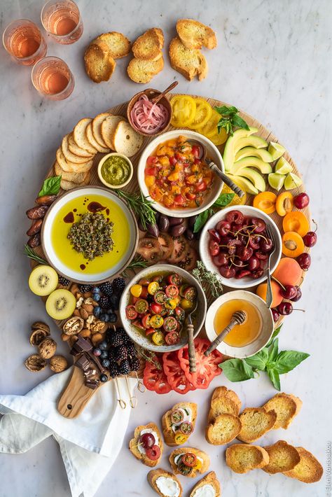 A wooden board with bruschetta toppings (pickled cherries, marinated tomatoes, apricot salsa, olive oil dip), fruit, nuts, avocado, pesto, tomatoes and chocolate. Around the board are some toasted slices of baguette. Everything is on a marble surface. Bruschetta Board Ideas, Apricot Salsa, Olive Oil With Herbs, Marinated Cherry Tomatoes, Bruschetta Board, Dipping Oil Recipe, Olive Oil Dip, Pickled Cherries, Marinated Tomatoes