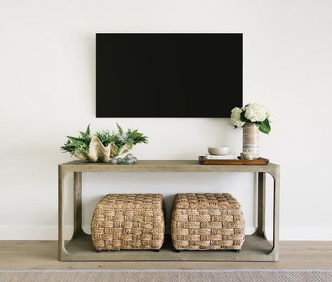 A flat panel television is mounted to a white wall above a Restoration Hardware Martens Console Table accented with a clamshell bowl, a wooden tray, and two seagrass poufs. Table Under Tv Decor, Table Under Mounted Tv, Console Table Under Mounted Tv, Table Under Tv, Console Table Under Tv, Living Room Console Table Decor, Under Tv Decor, Decor Under Tv, Mounted Tv Ideas Living Rooms