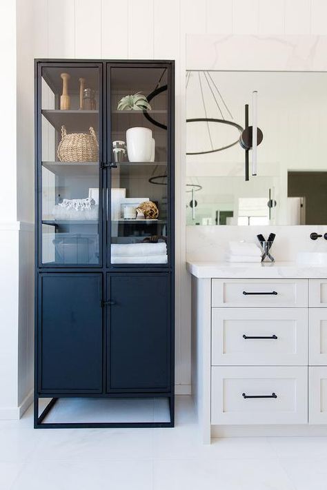 A freestanding black metal linen cabinet with glass doors is positioned beside a light gray washstand donning oil rubbed bronze pulls and a white quartz countertop fixed beneath a frameless vanity mirror. Bathroom Shelfie, Garage Studio, Bright Bathroom, Bathroom Linen Cabinet, Home Goods Store, Transitional Bathroom, Linen Cabinet, Interior Renovation, Glass Cabinet Doors