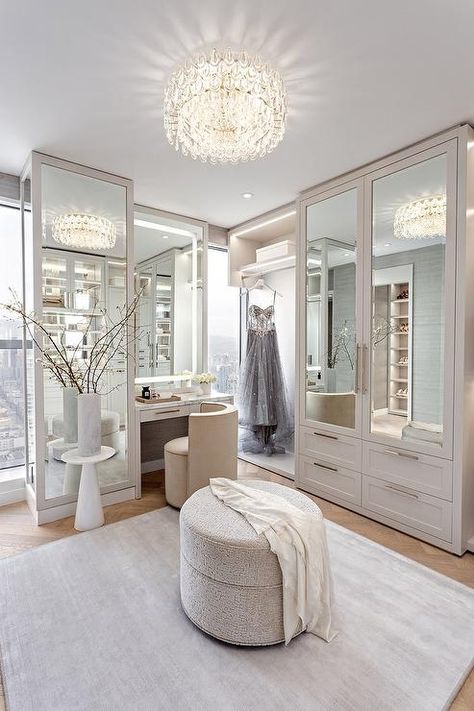 Glam light gray walk in closet illuminated by a crystal flush mount light features mirrored wardrobe cabinets, a round gray ottoman and a custom makeup vanity with cream velvet vanity chair. Gray Walk In Closet, Walk In Closet Design Ideas, Custom Makeup Vanity, Aesthetic Wardrobe Closet, Velvet Vanity Chair, Closet With Vanity, Organization Wardrobe, Gray Ottoman, Modern Wardrobe Design