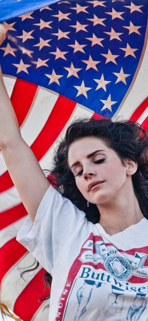 A white brunette, singer/songwriter Lana Del Rey,  stands with her eyes closed and wind blowing through her hair. She is holding the American flag above herself, letting it flow in the wind, and wearing a shirt that says Buttwiser, a spoof of Budweiser beer. Ride Lana Del Rey, Lana Del Rey Ride, Lana Del Rey Quotes, Iphone Wallpaper Music, Lana Del Rey Love, Lana Del Rey Lyrics, Brooklyn Baby, Vintage Americana, Lana Del Ray