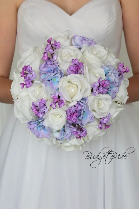 This is a round brides bouquet with white roses with jewel centers accented with purple/capri blue hydrangea and small purple lilacs. All of our wedding bouquets are made with artificial wedding flowers. Bouquet With Lilacs, White And Lavender Wedding, White Brides Bouquet, Blue Purple Wedding, Davids Bridal Gowns, Lilac Wedding Bouquet, Blush Bouquet Wedding, Purple Wedding Theme, Beautiful Wedding Flowers