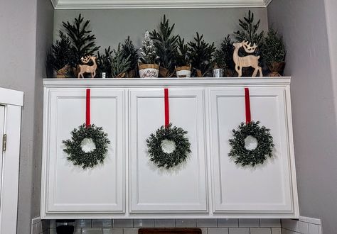 Christmas wreaths hanging on ribbon attached to kitchen cabinet doors. Different sizes of fake green Christmas trees on cabinet top with wooden reindeer for a refined but rustic holiday display. Cabinet Top Christmas Decorating Ideas, Garland On Top Of Cabinets, Upper Cabinet Christmas Decor, Garland Above Cabinets, Above The Cabinet Christmas Decor, Above Cupboard Christmas Decor, Christmas Village Above Kitchen Cabinets, Present Cabinets Christmas, Christmas Decor On Top Of Cabinets