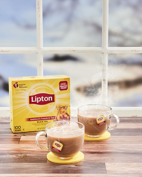 A box of Lipton Black Tea sits on a wooden table with two steaming mugs of black tea with milk sit on coasters next to it. The background is a window with an out of focus snowy landscape outside Hot Tea Recipes, Date Syrup, Lipton Tea, Black Tea Bags, Healthy Heart, Skim Milk, Bags Black, Hot Tea, Tea Bags