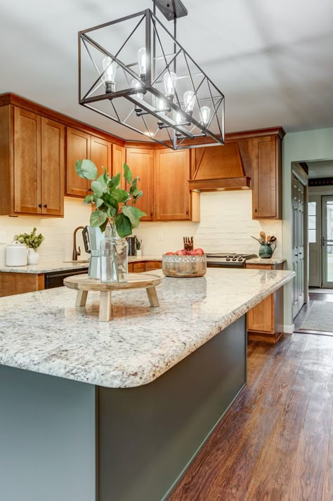 The contrasting warm and cool elements in this kitchen draw you in and makes you want to stay awhile. With white cabinets being so popular, these clients decided to go a different direction with brown maple wood wall cabinets. Adding a green island, light countertops, a white backsplash, and dark accents creates multiple textures, giving this kitchen dimension. The wall cabinets extend all the way to the large window, drawing in natural light, and the island offers bar seating. Island With Bar Seating, Island With Bar, Green Kitchen Island, Stained Kitchen Cabinets, Honey Oak Cabinets, Maple Kitchen Cabinets, Brown Kitchen Cabinets, Light Wood Cabinets, Green Island