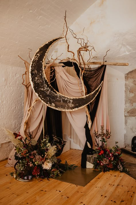 A large crescent moon in black and gold resin, in a geode style sits high in front of winding branches and drapes. Deep florals sit at the base of the ceremony arch, with a large golden candelabra with 5 candle sticks. Winter Solstice Wedding, Celestial Backdrop, Solstice Wedding, Celestial Wedding Theme, Starry Night Wedding, Festival Bride, Moon Wedding, Celestial Wedding, Dark Wedding