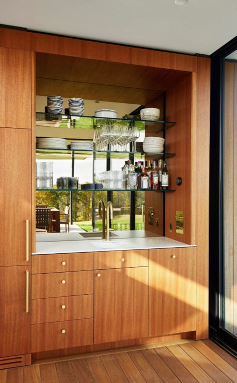 A wet bar that has rich, warm wooden cabinets with a reddish brown undertone. There is a white countertop with a gold faucet for the sink. There are three shelves above the sink with glassware, with a mirror backsplash against it. Modern Wet Bar, Modern Coffee Bar, Modern Basement Bar, Modern Home Bar Designs, Mid Century Modern Bar, Bedroom Inspirations Minimalist, Modern Home Bar, Modern Basement, Home Bar Design