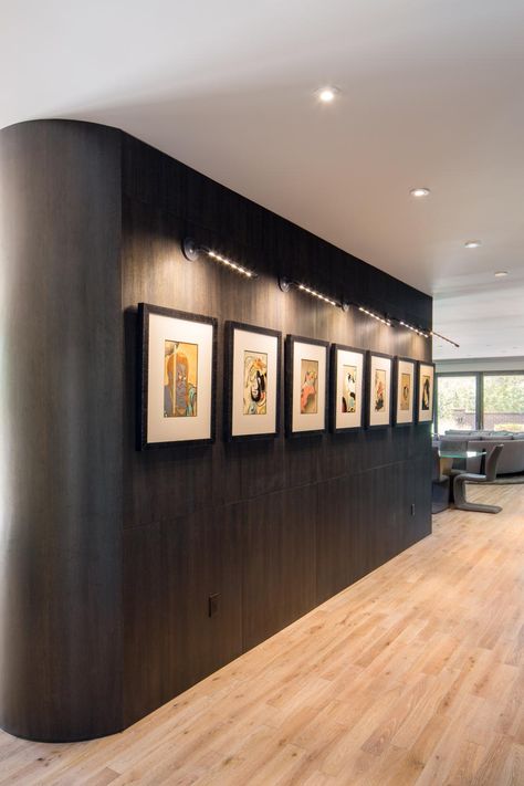 A dark wood wall contrasts against the pale hardwood floors in this open living room. Abstract art is lit by gallery lighting, creating the look of an exhibit with the homeowners' collection. Art Gallery Hallway, Hallway Opening, Art Gallery Lighting, Automobile Showroom, Art Showroom, Wall Frame Design, Wall Color Combination, Open Living Space, Art Galleries Design