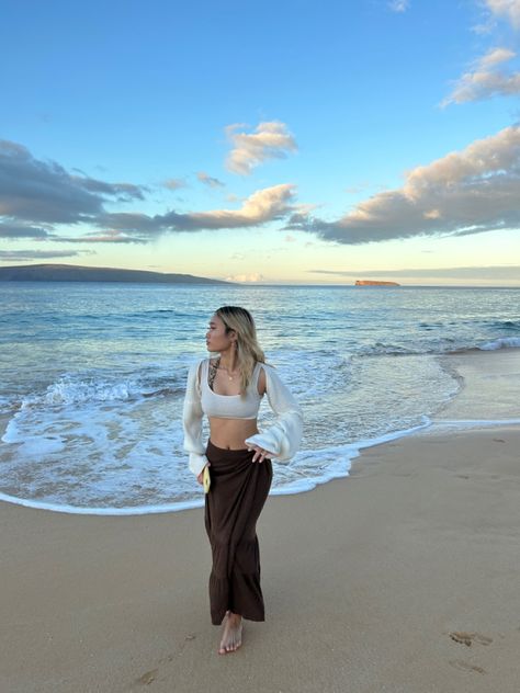 a girl walking horizontally towards the camera, facing away from the ocean with the waves and sunrise in the background. she’s wearing a long brown tiered skirt a short white tank top, and a white shrug or bolero. her hair is blonde and she’s holding a yellow phone case. White Shrug Outfit, Outfit Ideas Beach, Brown Maxi Skirts, White Shrug, Beach Girl Aesthetic, Beach Fits, Brown Skirts, Brown Outfit, Beach View