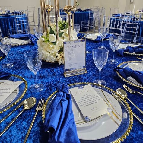 Behold the enchanting wedding banquet, resplendent in all its glory! The regal blue velvet tablecloth drapes gracefully, captivating hearts at first sight. Nestled upon clear round charger plates, royal blue satin dinner napkins exude opulence. Delicate white wax candles flicker inside long, transparent candleholders, casting a romantic glow. A small bouquet of faux white silk roses graces each table, adding a touch of everlasting beauty. Royal Blue Centerpieces Quinceanera, Blue Dinner Party, Royal Blue Centerpieces, Centerpieces Quinceanera, Velvet Tablecloth, Event Decor Ideas, Party Decorations Table, Group Dinner, Candles Flowers