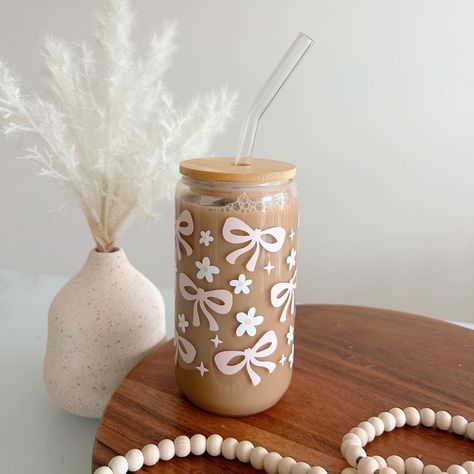 Bows and Daisys Tumbler with Straw - 16oz Celebrate your love for coffee with this glass tumbler! This 16oz tumbler is perfect for every caffeine lover out there. This glass tumbler comes with a natural wooden lid and a sleek, reusable glass straw, making it both a chic and sustainable choice for your drinkware collection. Perfect for iced coffee, smoothies, or any cold beverage, this tumbler is a great companion for busy days, whether at home or on the go. It also makes a thoughtful gift for all the special people in your life! Product Details: *Capacity: 16oz *Material: Glass with a wooden lid *Includes: Reusable glass straw *Design: Daisys and bows *Ideal for cold beverages *Eco-friendly and reusable Care Instructions: *Hand wash only *Do not microwave  *Do not put in dishwasher Sip in Iced Coffee To Go Cup, Cute Coffee Glass Cups, Cute Glass Coffee Cups, Glass Tumbler With Straw, Iced Coffee Cups, Painting Glass Jars, Straw Design, Trendy Water Bottles, Cute Coffee Cups