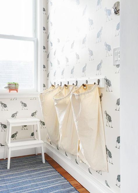 Mounted on a wall covered in  Beware The Moon Ostrich Wallpaper, three hampers hang on hooks on a wall adjacent to a window positioned above a white step stool placed on a black and white rug. Ostrich Wallpaper, Laundry Room Hamper, Kitchen Wall Covering, Laundry Rooms Ideas, Transitional Laundry Room, Laundry Shelves, Laundry Room Closet, Laundry Sorter, Laundry Room Renovation