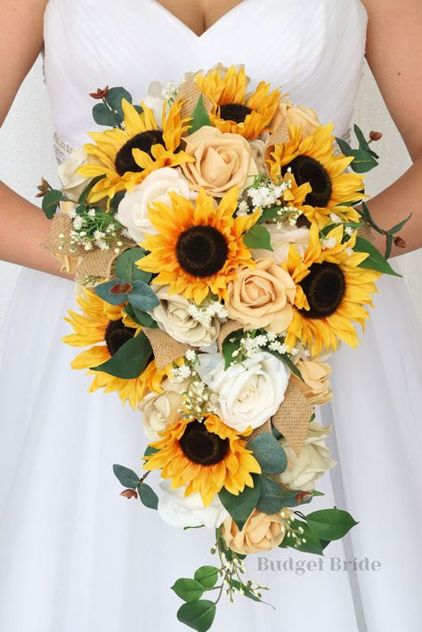 This striking cascading brides’ bouquet is made with an elegant combination of beige, champagne, and sunflowers to create a beautiful amount of depth and contrast within this bouquet. The sunflowers and roses alongside the variety of mixed foliage and filler make this bouquet a stand out. As always, our bouquets are wonderful for those who are allergic to flowers and pollen, and have the added bonus of not attracting any bees or bugs. All of our bouquets are wired by hand, so that you can rest a Western Wedding Bouquets With Sunflowers, Sun Flower Wedding Theme Bridesmaid Dresses, White Roses And Sunflower Bouquet, Summer Sunflower Bouquet, Bride Bouquets Sunflowers, Sunflower Hydrangea Bouquet, Sunflower Bride Bouquets, White Sunflower Bouquet, Sunflower Bouquet Aesthetic