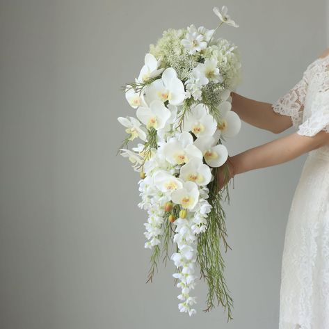 💐Product Description: This exquisite bridal bouquet showcases a stunning arrangement of white flowers, meticulously crafted to create a cascading effect. The bouquet features elegant white orchids as the focal point, complemented by smaller white blooms and delicate greenery. The design creates a waterfall-like appearance, adding a touch of sophistication and grace to any bridal ensemble. The flowers are thoughtfully arranged to ensure a balanced and harmonious look, with the cascading elements Wedding Flowers For Virgin Mary, White Flower Wedding Bouquet Dui, Silk Vs Real Flowers For Wedding, White Flowers And Greenery Bridal Bouquet, Orchid Bouquet Wedding, Lily Bridal Bouquet, Teardrop Bouquet, Cascading Bridal Bouquets, Cascading Wedding Bouquets