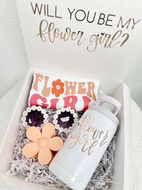 Present your flower girl with a delightful proposal box that will surely make her smile. Inside, she'll discover a charmingly colorful flower girl t-shirt, adorned with whimsical designs to celebrate her special role. Accompanying this are adorable flower daisy sunglasses, perfect for adding a touch of playful flair to her ensemble. To make her feel even more special, include a personalized sippy cup or mini tumbler in her favorite color, ensuring she can stay refreshed in style throughout the c Bridesmaid Proposal Box Outside, Wedding Favors For Bridesmaids, Wedding Bridesmaid Ask Proposals, Cute Bridesmaid Proposal Boxes, Braidmaids Proposal Boxes, Cozy Bridesmaid Proposal, Flowergirl Proposal Gift, Flower Boy Proposal, Wedding Proposal Boxes