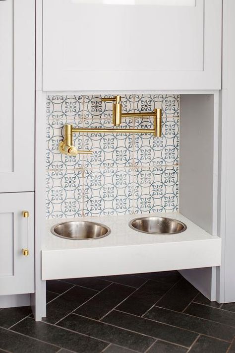 A brass swing arm pot filler is mounted to white and blue mosaic backsplash tiles over a built-in dog food bowl fixed above black herringbone floor tiles and between light gray-blue cabinets with glass and brass pulls. Black Herringbone Floor, Dog Station, Dog Food Station, Transitional Laundry Room, Mudroom Flooring, Dog Feeding Station, Grey Floor Tiles, Hydration Station, Bathroom Cabinetry