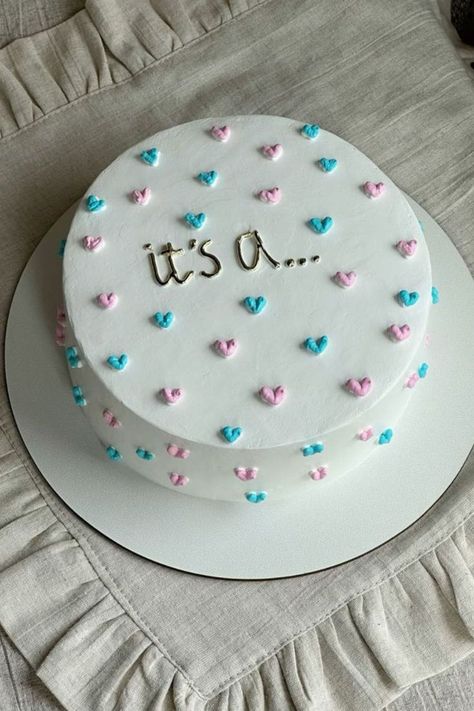 A gender reveal cake with white frosting and pink and blue hearts, with the text "It’s a..." written on top. Simple Reveal Ideas, Gender Reveal Lunchbox Cake, Gender Reveal Cake Diy Easy, Gender Reveal Cake Sayings, Gender Reveal Cutout Cookies, Easy Cute Gender Reveal Ideas, Cheesecake Gender Reveal, Ways To Do Gender Reveal, Dessert For Gender Reveal Party