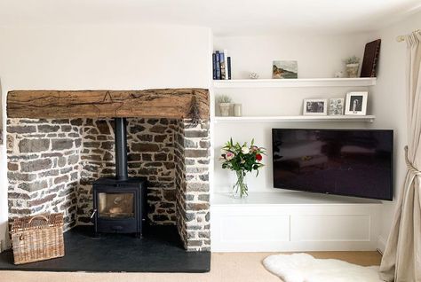 Black wood burning stove is placed inside a stone fireplace with black hearth and rustic wood mantel. A flat panel TV is positioned inside a white built-in media unit next to the fireplace. White Fireplace Mantels, Painted Stone Fireplace, Grey Stone Fireplace, White Stone Fireplaces, Woodburning Stove Fireplace, Fireplace Facing, Fireplace Feature Wall, Stacked Stone Fireplaces, Living Room Ceiling Fan