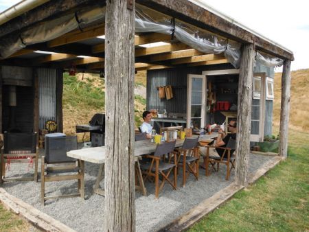 A covered outdoor kitchen area, with Nick and Kai sitting at a long table. Outdoor Glamping Kitchen, Glamping Kitchen Outdoor, Glamping Kitchen, Outdoor Kitchen Area, Bbq Shed, Covered Outdoor Kitchens, Rustic Outdoor Kitchens, Outdoor Camping Kitchen, Outdoor Grill Station