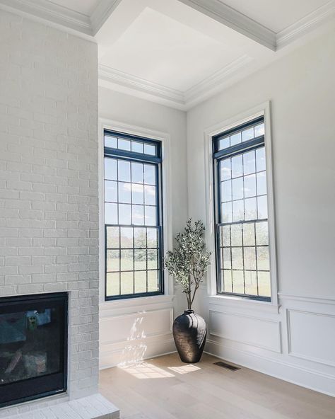 This bright living room features large windows with modern black trim and black grids. The size of the windows lets ample natural light into the space, illuminating the beautiful wainscoting and coffered ceiling. The modern white brick fireplace is a cozy addition to this transitional space. Modern Window Trim Ideas, Modern Black Windows, Modern Window Trim, Black Trim Interior, Window Trim Ideas, Window Trim Styles, White Window Trim, Nailhead Furniture, Black Window Trims