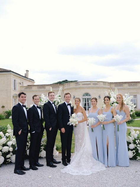 Bridal party photo with bride, groom, bridesmaids in light blue dresses, and groomsmen in black suits with black bow ties, set against the elegant Chateau Gassies. Black Tux With Light Blue Tie, Bridesmaid Dresses Groomsmen Suits, Bridal Party Color Palette, Black Suit With Blue Tie, Black And Light Blue Wedding, Light Blue Bridal Party, French Blue Wedding Color Palettes, Blue Bridesmaid Dresses With Groomsmen, Chateau Gassies