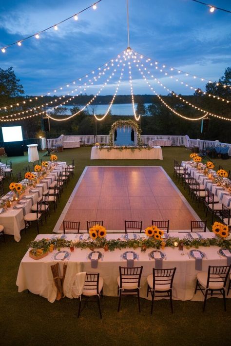 An outdoor wedding reception setup with a dance floor in the center, surrounded by tables decorated with sunflowers and greenery. String lights hang above, creating a romantic atmosphere. A scenic view of water and trees is in the background. Small Modern Wedding Ideas, Outdoor Diy Wedding Reception, Wedding Ideas Minimal, Wedding Inspo Romantic Small, Good Wedding Ideas, Cute Intimate Wedding Ideas, Small Wedding Reception Ideas Outdoor, Backyard Wedding Ceremony Small, Small Outdoor Wedding Ideas Backyards