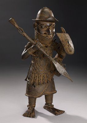 This bronze figure represents a warrior chief, and would have been placed on a shrine to honor an Oba, or king, accompanied by a memorial head and tusk, bells, and other ceremonial items. The figure’s garments and accoutrements all make reference to his status as a warrior with royal associations. He holds a shield in one hand and a ceremonial spear in the other, and the series of small bells that hang from his garment are indications of his status within the warrior class. The leopard – r... Oyo Empire, African Armor, Benin Empire, Medieval Africa, Ancient Mali, Benin Bronzes, Benin Art, Benin Bronze, African Artifacts