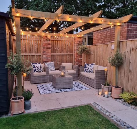 A gray wicker patio set is arranged on a white and black rug laid over beige stone pavers. The patio set is covered by a light wood pergola lit by black festoon lights. Make pergolas cozier with greenery, garden lights, and black lantern dÃ©cor. Fenced In Pergola, Backyard Vision Board, Garden Patio With Pergola, Garden Ideas Pergola, Pergola In Yard, Garden Design With Pergola, Patio With Fairy Lights, Back Garden Design Ideas, Pergola Next To House