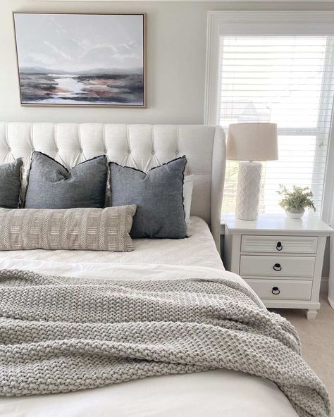 This bedroom features a white tufted bed pushed against the off-white wall underneath a landscape picture. A white bedspread covers the bed on which a light grey coverlet has been tossed. Dark grey pillows and a beige bolster have been placed at the head of the bed. A white bedstand sits on the beige carpet in front of a bright window. Bedroom Ideas Cream Carpet, Grey Bed Pillows, Cream And White Layered Bedding, Cream And Grey Bed, Bed Blanket Arrangement, School House White Bedroom, Gray And Beige Bedding, Grey And White Bedding Ideas, White Bed With Colorful Pillows