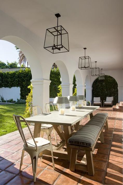 Chic covered patio with arched doorways boasts a tow of iron and glass lanterns illuminating a concrete and wood outdoor dining table lined with white Tolix Chairs and a sawhorse bench atop a terracotta floor. Patio Arches Spanish Style, Arched Outdoor Patio, Arched Patio Cover, Patio Archway, Patio With Arches, Arched Patio, Terracotta Outdoor Patio, Patio Arch, Terracotta Patio