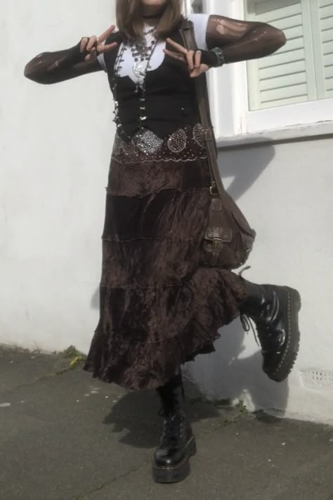 A mid-size person/girl wearing an oversized white fairy crop top, a black waistcoat over it, a black ripped tights top under it, a long dark brown skirt, ripped rose-patterned tights under it, black platform dr. Martens and multiple crystal rings and bracelets. 	The person is also wearing multiple silver necklaces; one in form of a fairy and one in form of a moon, a brown rosary-style and a green bead necklace and a black lace choker. They also have a brown handbag with them. Brown Goth Aesthetic, Maxi Skirt Outfit Grunge, Brown Goth Outfit, Crow Core Aesthetic Outfits, Crow Core Outfit, Cottage Goth Fashion, Steampunk Aesthetic Outfit, Dark Cottage Core Outfits, Fit Aesthetic Outfit