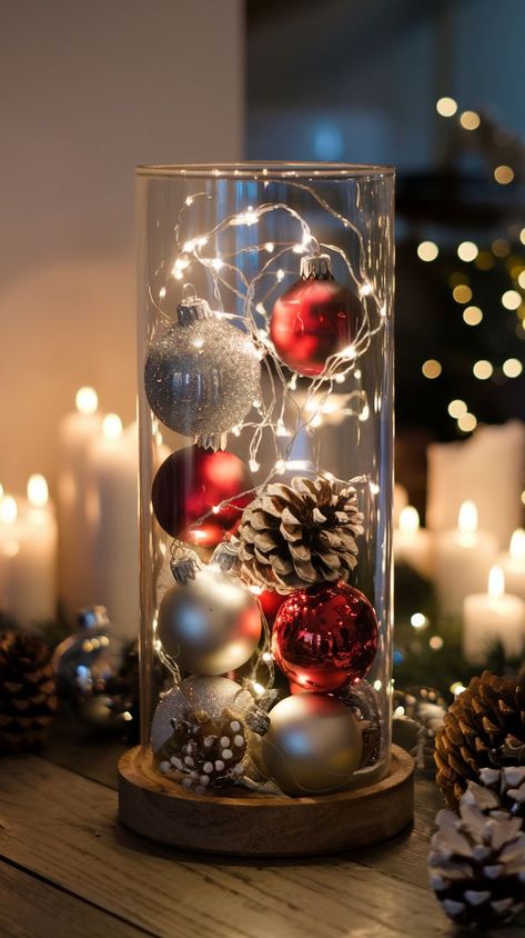 A glass cylinder filled with red and silver Christmas ornaments, pinecones, and string lights creates a cozy, festive ambiance. Lit candles in the background. Christmas Wedding Centerpieces Diy, Ornament Centerpieces, Glass Vase Christmas Decor, Christmas Dinner Table Centerpieces, Cheap Table Decor, Table Scape Ideas, Table Decor For Christmas, Christmas Centre Pieces, Christmas Party Centerpieces