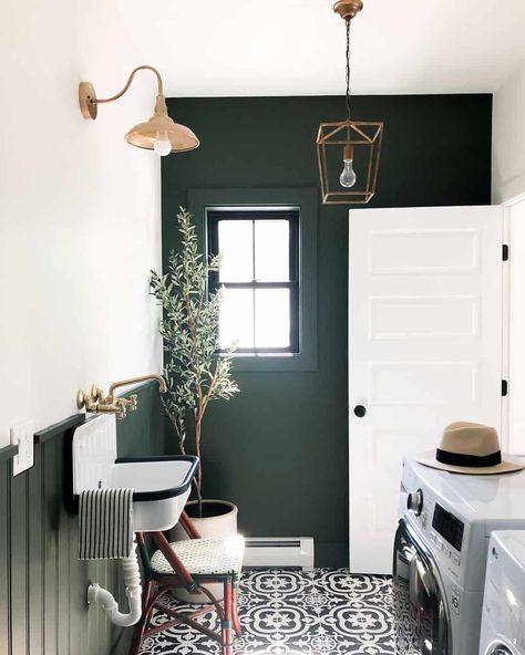 This modern laundry room displays dark wood wainscoting and a feature wall that contrasts with light white painted walls. Gold accents are found in the faucet above a vintage sink, as well as the light fixtures that illuminate the gray and white patterned tile floor. Laundry Room Paint Color, Laundry Room Paint, Wood Wainscoting, Wall Mount Kitchen Faucet, White Laundry Rooms, Vintage Sink, Green Laundry, Modern Laundry, Dark Green Walls