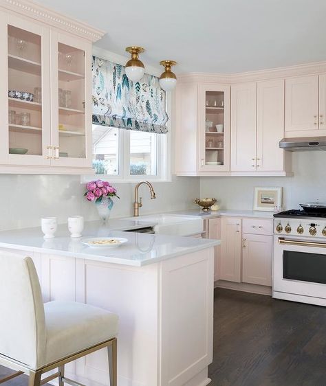 Contemporary kitchen features soft pink cabinets with glass and brass pulls, a white farm sink that boasts a brass gooseneck faucet under a window with a blue and white roman shade, lit by Clark flush mount lights, a pink kitchen peninsula topped in white quartz with white leather counter stools, a white and gold stove and dark wooden floors. White Cabinets Pink Walls, Kitchen Peninsula Window, Soft Pink Kitchen Cabinets, Blush Pink Cabinets, Pink And White Kitchen Cabinets, Pink Kitchen Sink, Light Pink Cabinets Kitchen, Blush Pink Kitchen Cabinets, Light Pink Cabinets