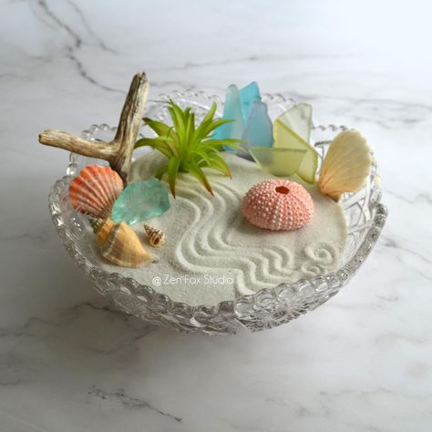 Add some coastal vibes to your space with this Beachy Zen Garden display in a Vintage Imperial Glass Bowl! This one of a kind sand garden sits inside an ornate vintage glass bowl. The air plant is artificial, no maintenance needed. Try adding your own treasures to personalize it!  If you need to relax, rake or trace designs in the sand and rearrange the accessories. Great for display on any desk, bookshelf, or table top.  Includes everything pictured: Imperial Glass Bowl, White Sand, Rake, Bamboo Stylus, 8 Pieces of Sea Glass, 4 Sea Shells, Sea Urchin, Driftwood, Faux Air Plant Base Diameter: 7.75" ★See all of the Zen Gardens available in my shop: https://www.etsy.com/shop/ZenFoxStudio Zen Garden Desk, Sand Boxes, Glass Bowl Centerpieces, Zen Sand, Sand Garden, Shell Mermaid, Diy Succulent Terrarium, Succulent Bowls, Garden Centerpiece