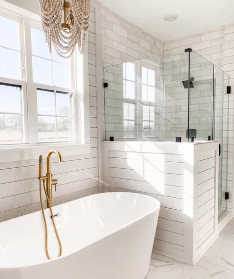 A minimalist white bathroom design showcases a clear glass shower with white subway tile and white flooring enclosed within. A white bathtub with a brass faucet rests outside the shower below a white window that fills the space with bright natural light. A beaded chandelier hangs over the tub and light tile flooring runs throughout...   Image: sixandstonehome Bathrooms With Soaker Tubs, Bathroom Soaker Tub, White Minimalist Bathroom, Shower With Glass Door, Soaker Tubs, Clear Glass Shower Door, White Subway Tile Shower, Tub Lighting, Luxurious Bathtubs