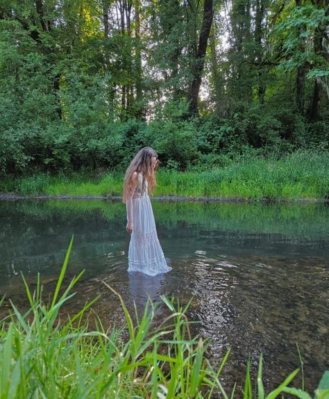 A girl in a white dress stands in the middle of the river by a forest. It appears she is floating White Dress Forest, White Dress In Water, Creepy Cottagecore, River Shoot Photography, River Photoshoot Ideas, White Dress River Photoshoot, River Photoshoot Models, Scarlett Aesthetic, Senior Pictures River Water